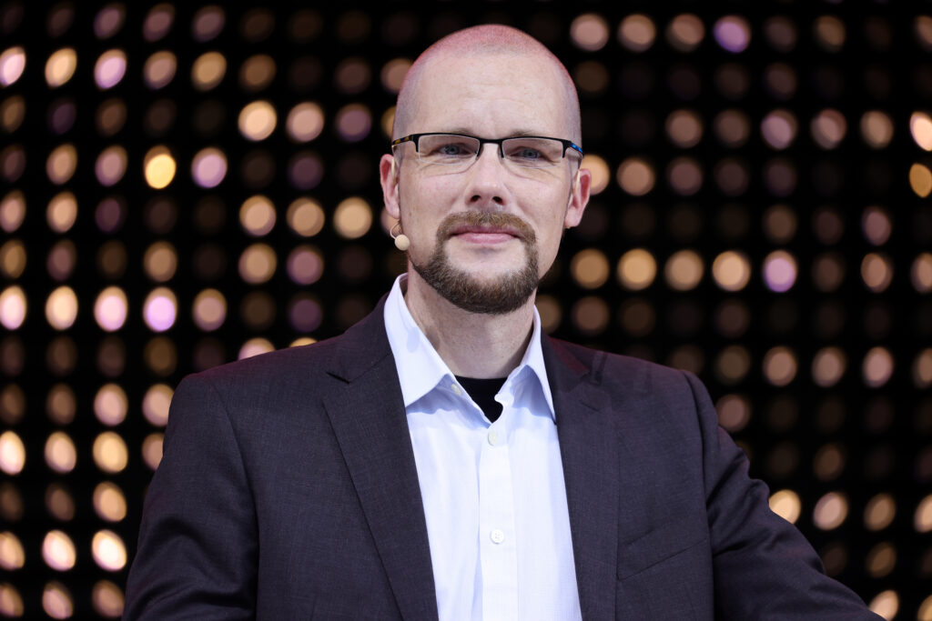 Jonas Andrulis mit schwarzem Sakko und weißem Hemd sowie Brille lächelt in die Kamera