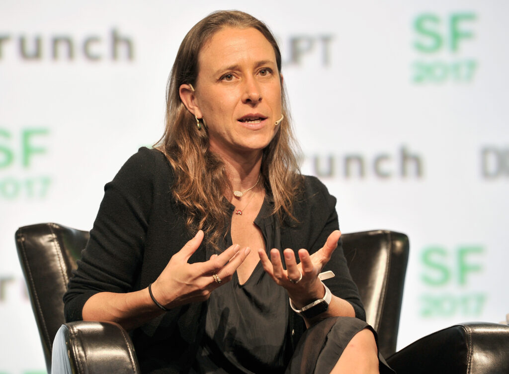 Anne Wojcicki sitzt mit einem schwarzen Kostüm auf einer Bühne und gestikuliert mit den Händen