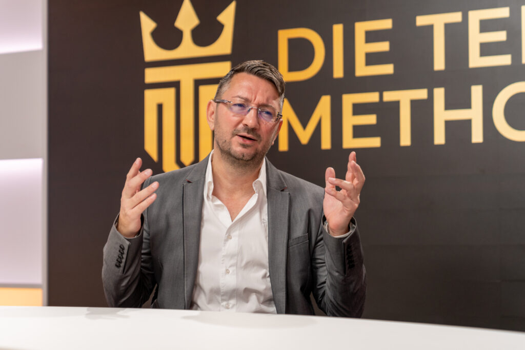 Walter Temmer sitzt gestikulierend vor einer Wand mit der Aufschrift 