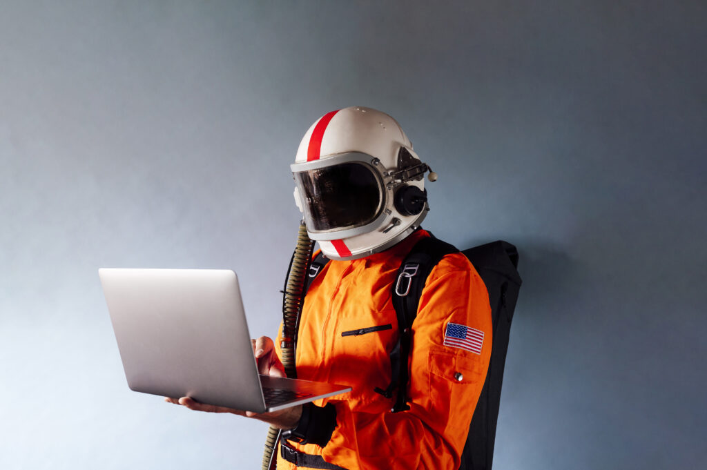 Ein Mensch in einem orangen Raumanzug mit weißem Helm der in der Mitte einen roten Streifen hat. Der Astronaut hält ein Notebook in seiner Hand