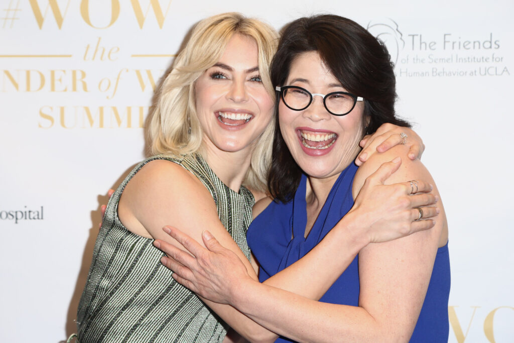 Wendy Suzuki umarmt eine Frau mit blonden Haaren. Suzuki trägt ein blaues Kleid ohne Ärmel und eine schwarze Brille. Beide lachen in die Kamera
