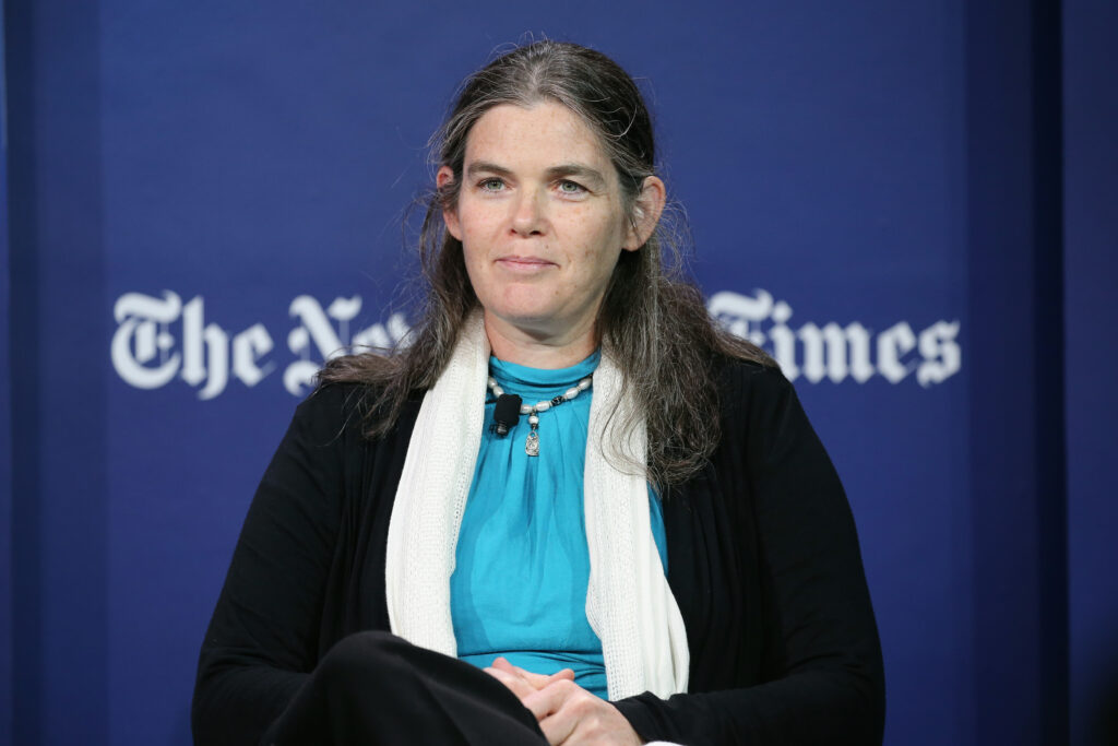 Daphne Koller mit einem türkisen T-Shirt und schwarzem Sakko sitzt auf einer Bühne. Hinter ihr das Logo der New York Times
