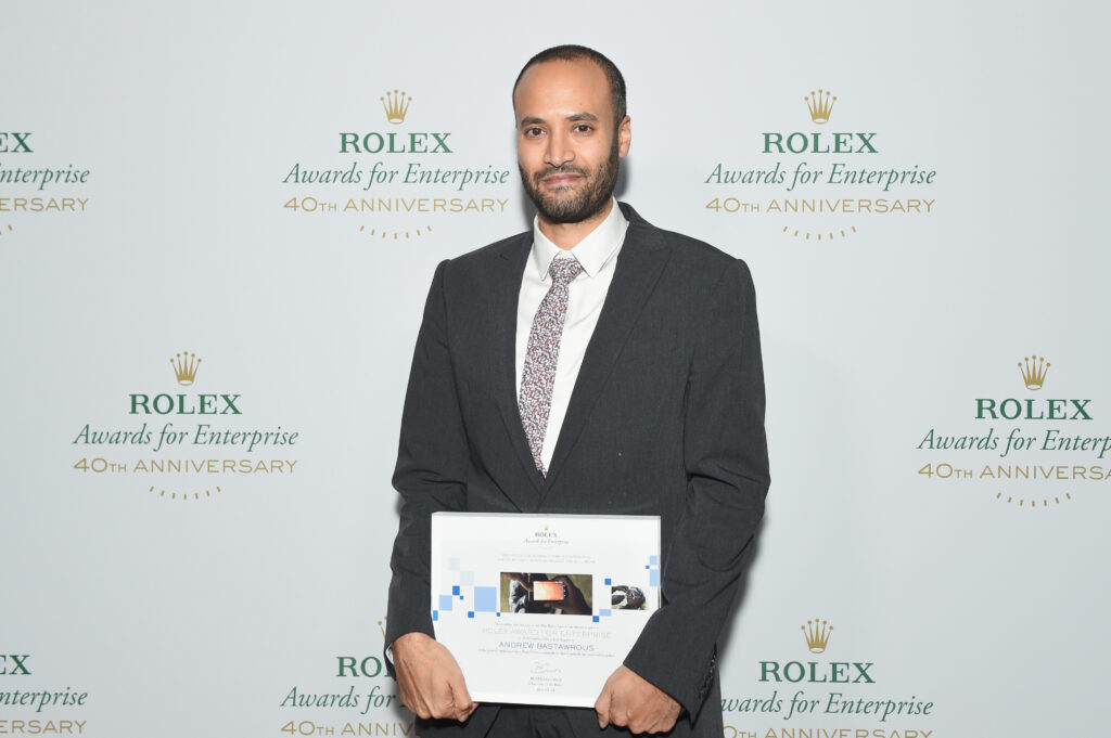 Andrew Bastawrous im Anzug blickt i die Kamera. Dahinter das Logo von Rolex Awards for Enterprise