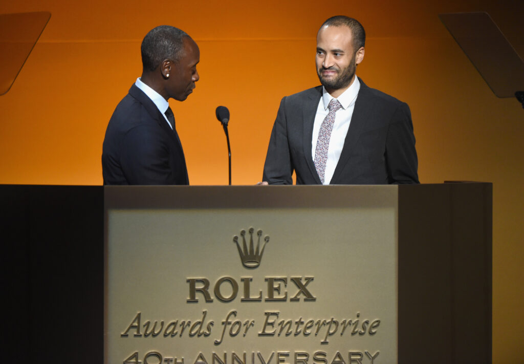Andrew Bastawrous steht mit einem Anzug neben einem anderen Mann auf der Bühne. Davor das Logo von Rolex Awards for Enterprise