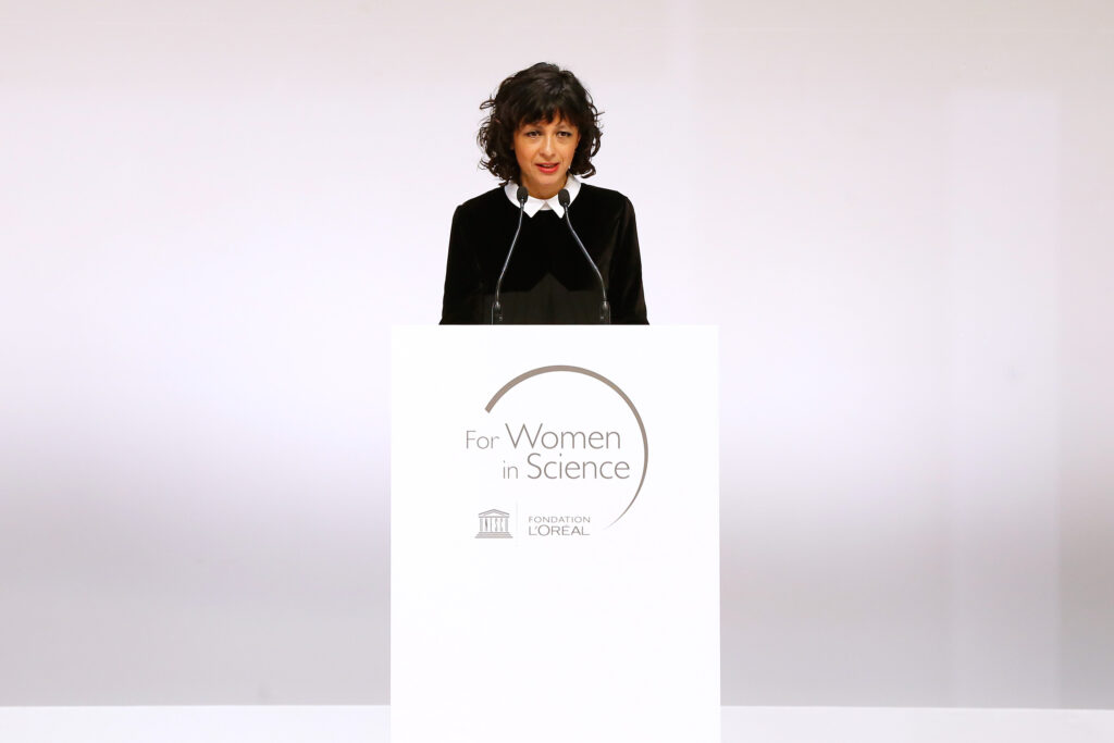 Emmanuelle Charpentier mit schwarzem Oberteil und weißer Bluse steht an einem Podest mit der Aufschrift: For Women in Science