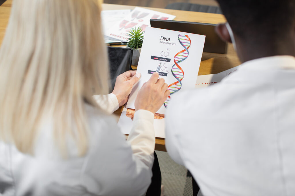 Eine Frau mit blonden Haaren und ein Mann von hinten aufgenommen. Die Frau hält einen Kugelschreiber auf eine DNA-Analyse