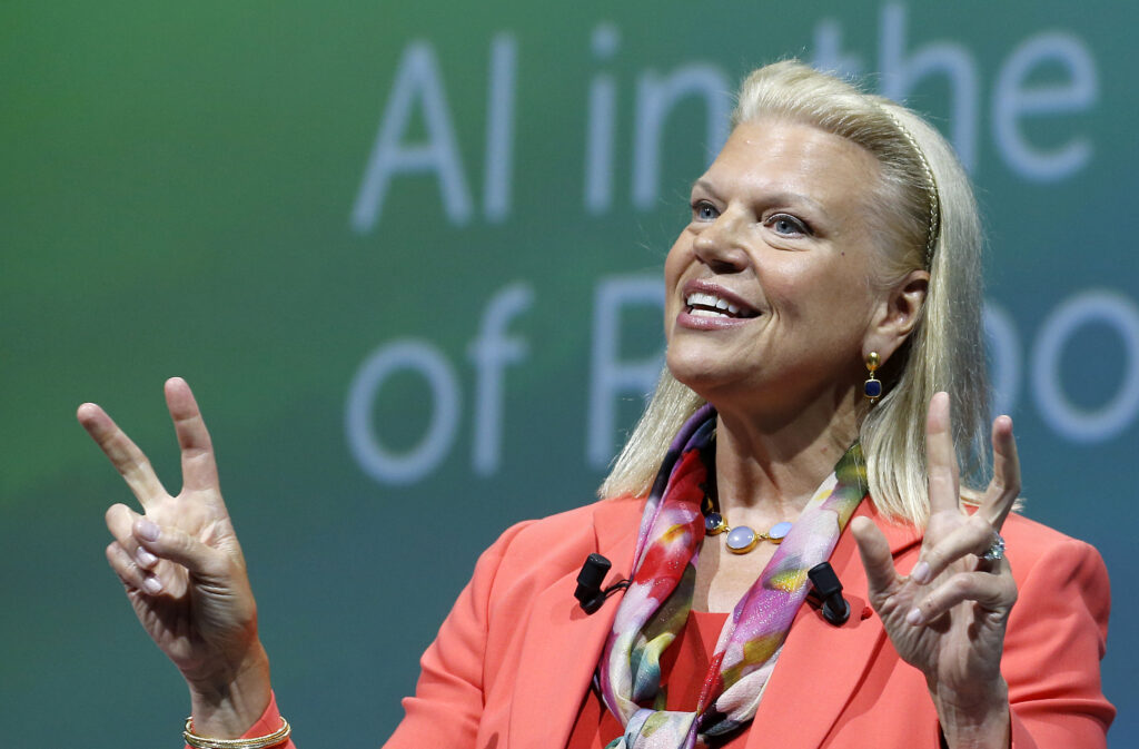 Ginni Rometty mit orangem Sakko hält beide Hände in die Höhe und spricht zu einem Publikum
