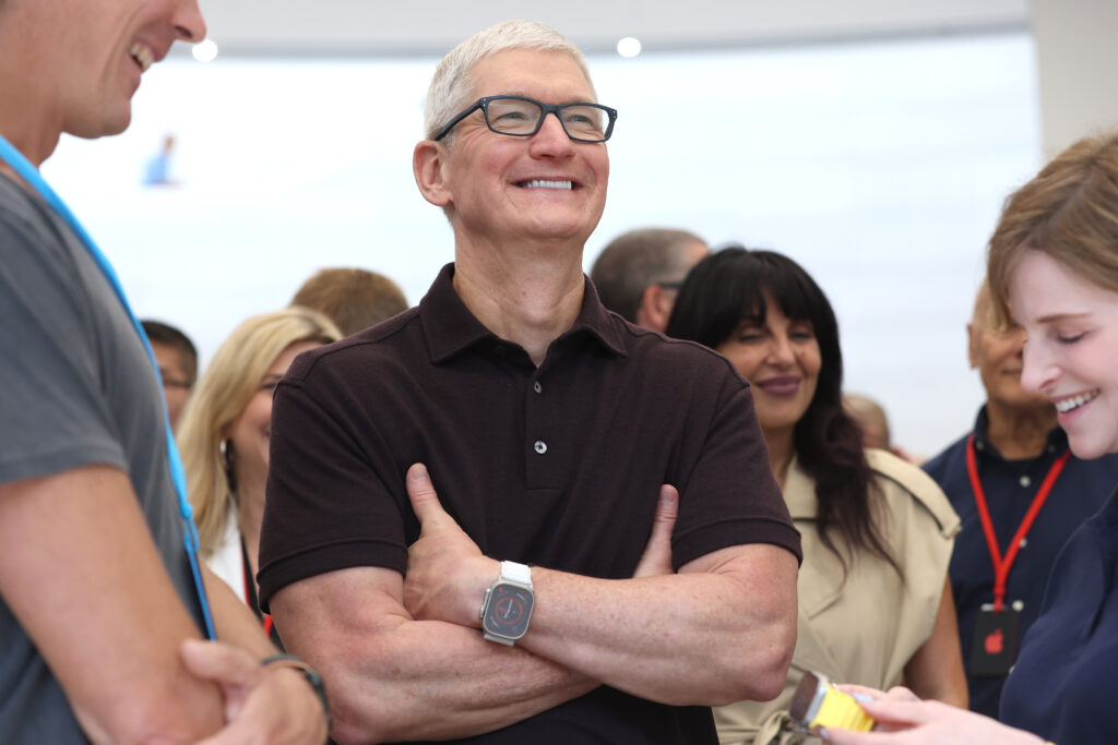 Tim Cook mit verschränkten Armen und Brille trägt ein schwarzes T-Shirt und lächelt in das Publikum