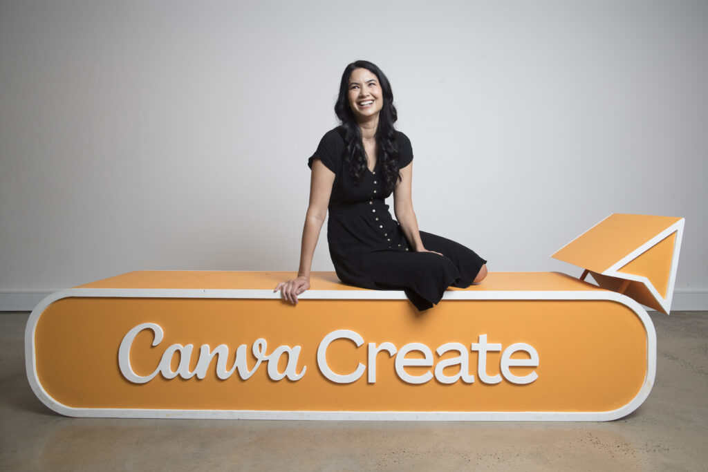 Melanie Perkins sitzt mit einem schwarzen Kleid auf dem Logo in orange von Canva Create