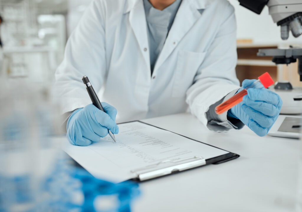 Forscher mit blauen Hygienehandschuhen hält ein rotes Röhrchen mit Flüssigkeit und macht sich mit einem Kugelschreiber Notizen auf einem Block 